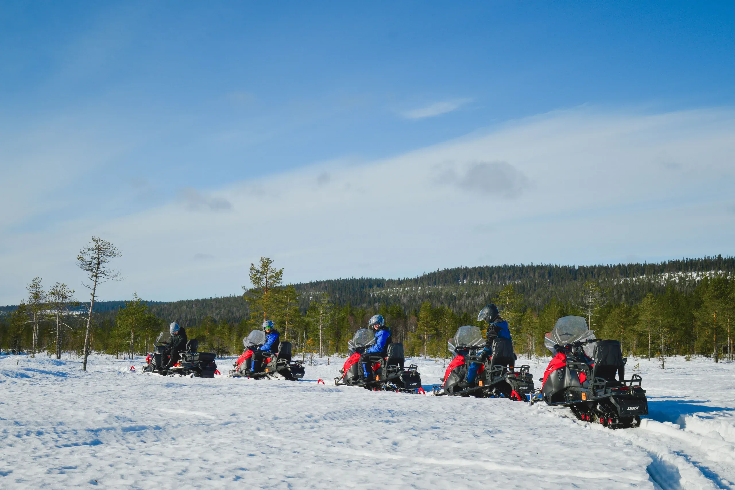 Five snowmobiles in a line across an open snowfield, riders in helmets, with a low forested hill and a wide blue sky behind them