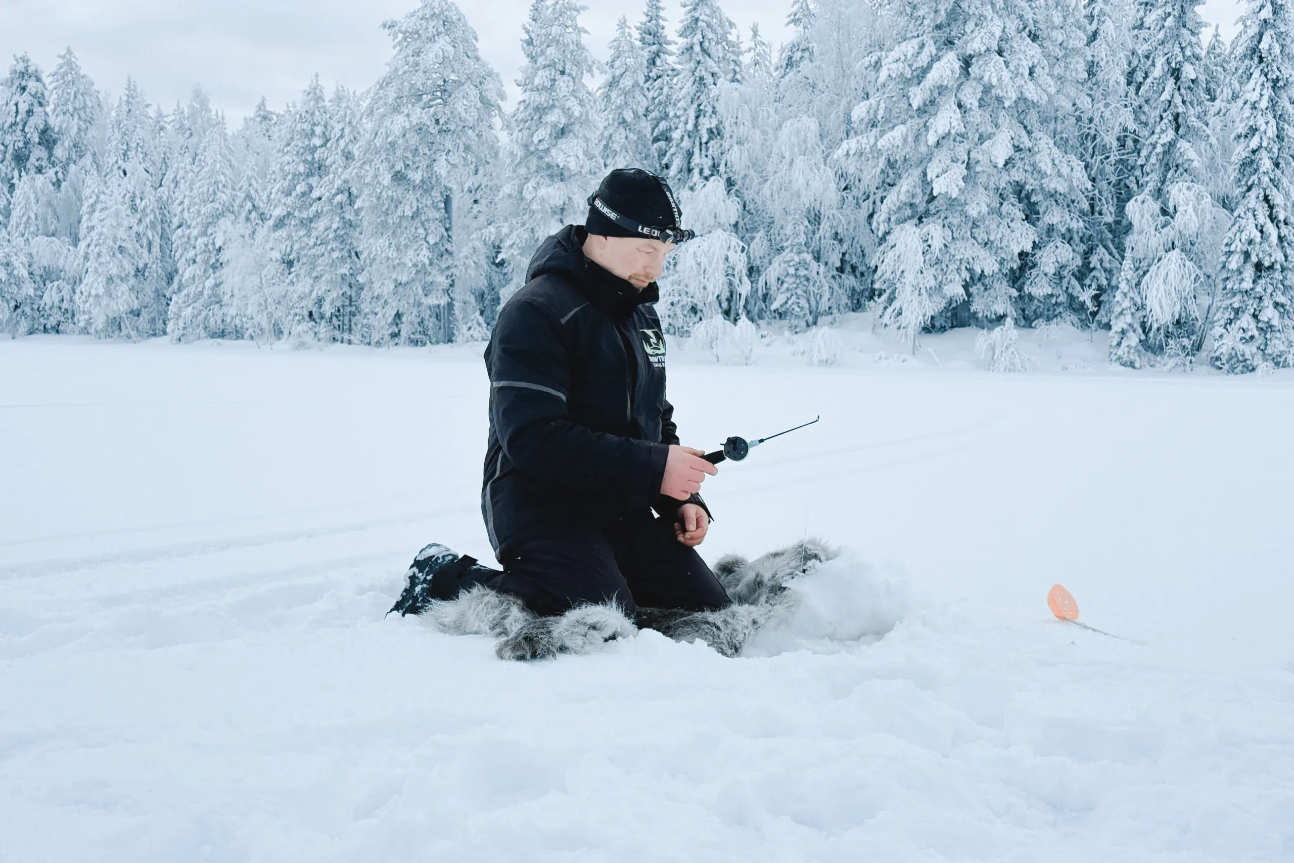 A guide in a black Snowtrail jacket and headlamp kneeling on a reindeer hide on a frozen lake, holding a short jigging rod beside a drilled hole, with an orange ice scoop on the snow and snow-laden spruce forest behind