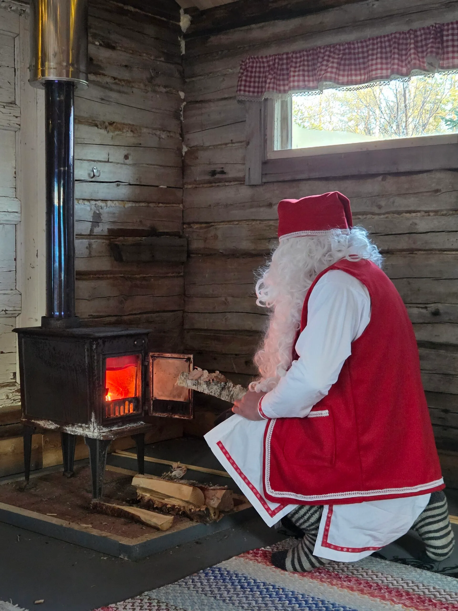 Santa Claus in a red waistcoat and hat, seen from behind, kneeling to feed birch logs into a glowing wood stove in a log cabin