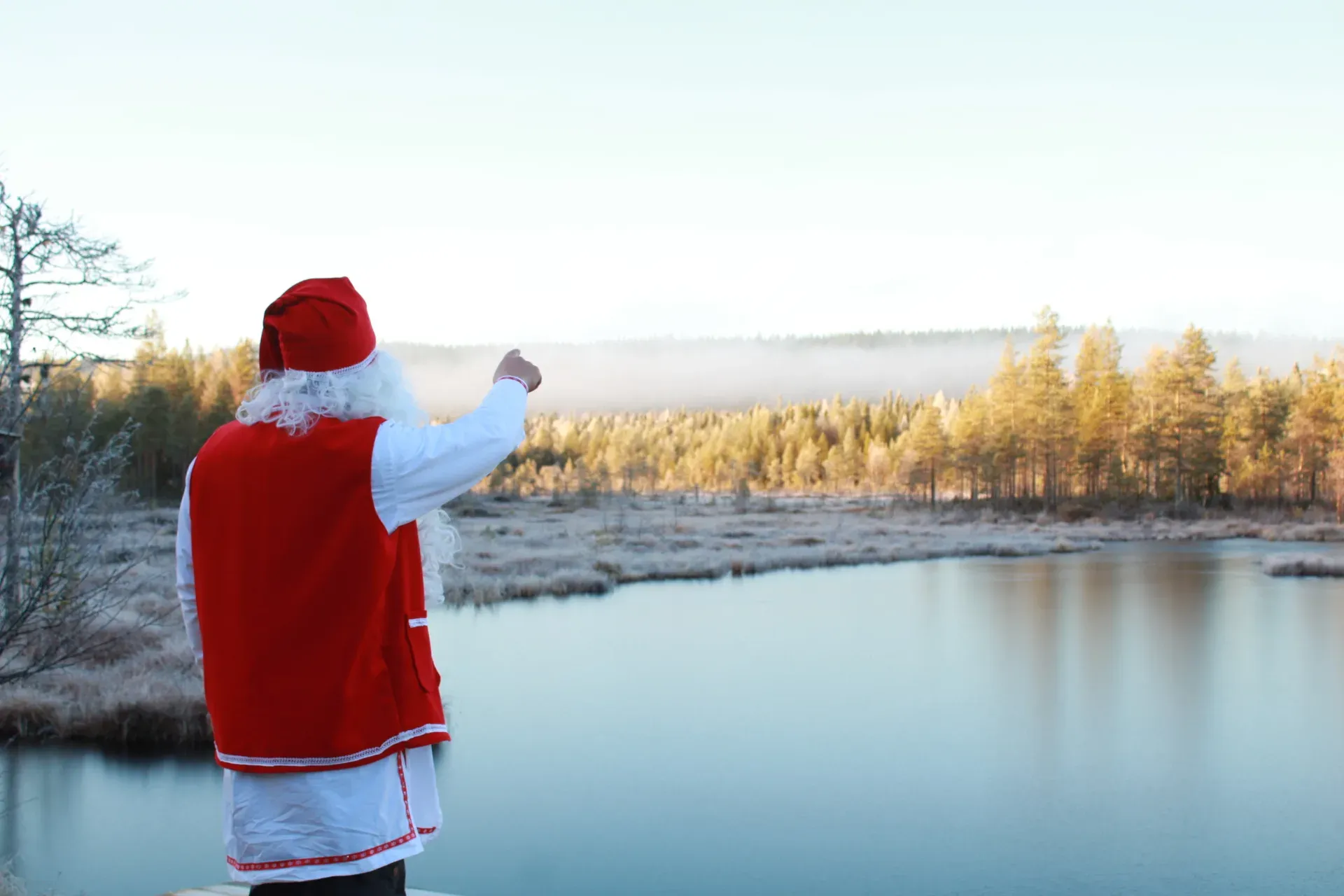Santa Claus in a red waistcoat and hat, seen from behind, pointing across a still lake towards frosted autumn pines lit gold by low sun, with mist lying over the far shore