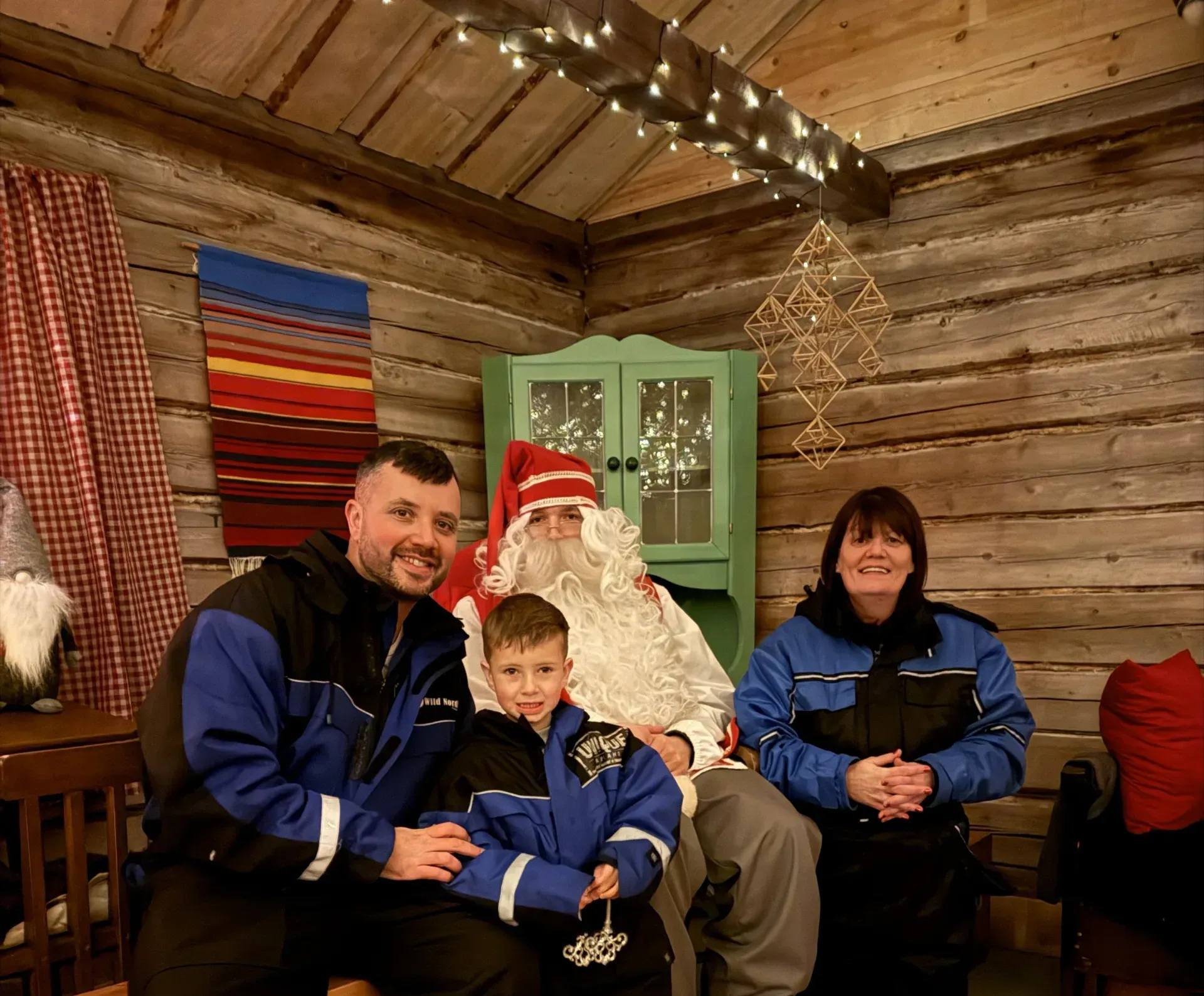 A man, a woman and a young boy in blue winter overalls sitting beside Santa Claus in a log cabin hung with fairy lights, a woven wall hanging and a green painted cupboard behind them