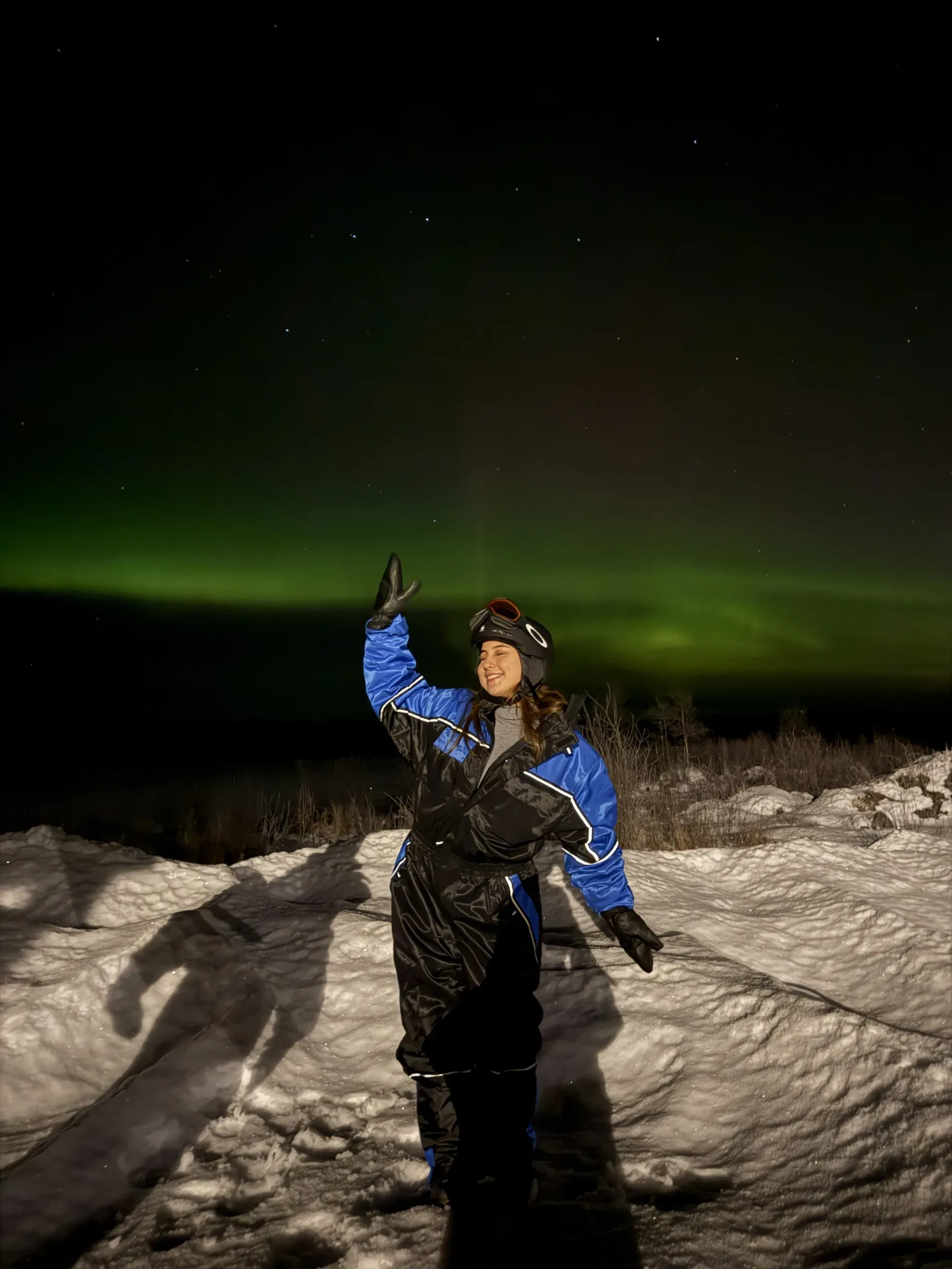 A woman in a blue and black snowmobile overall with goggles on her helmet waving on a snowbank at night, a band of green aurora low on the horizon behind her