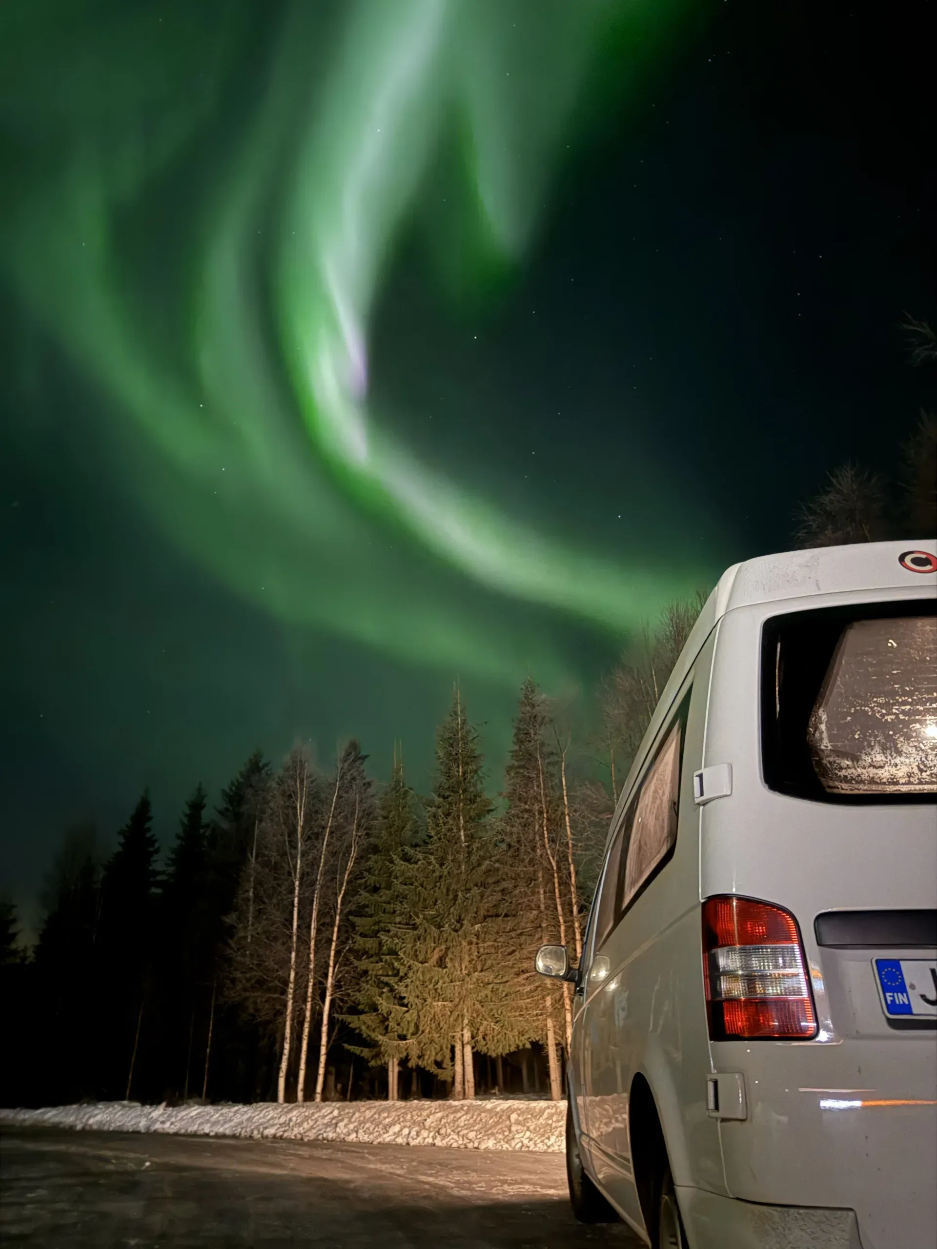A bright green aurora arcing over birch and spruce at night, beside the frosted rear window of a parked white minibus