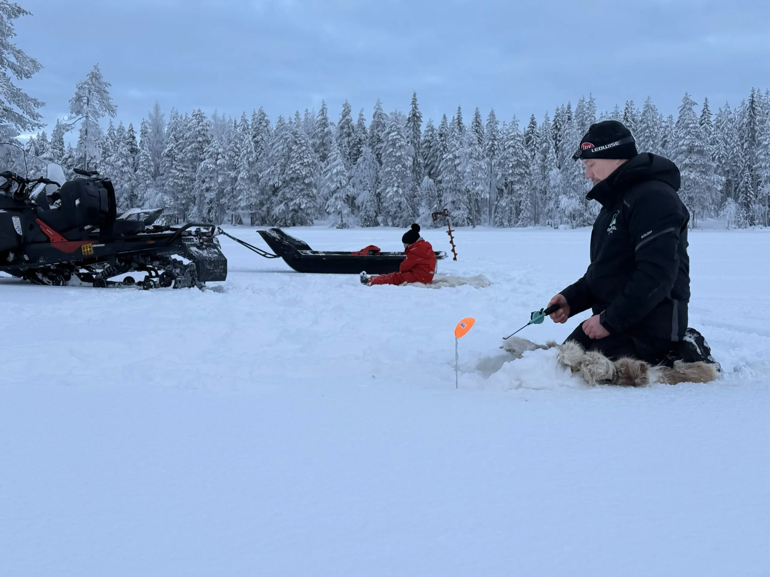 A guide kneeling on a snow-covered frozen lake with a short jigging rod beside a marker flag, a snowmobile and towed sleigh parked behind him and snow-laden spruce along the shore