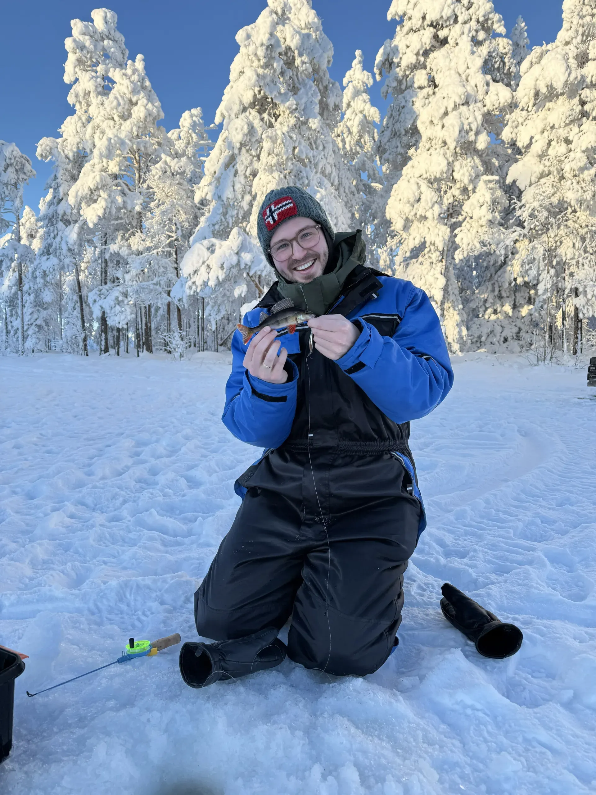 A smiling man in a blue and black winter overall and a knitted hat kneeling in snow on a frozen lake, holding up a small perch in both hands, with snow-covered forest behind