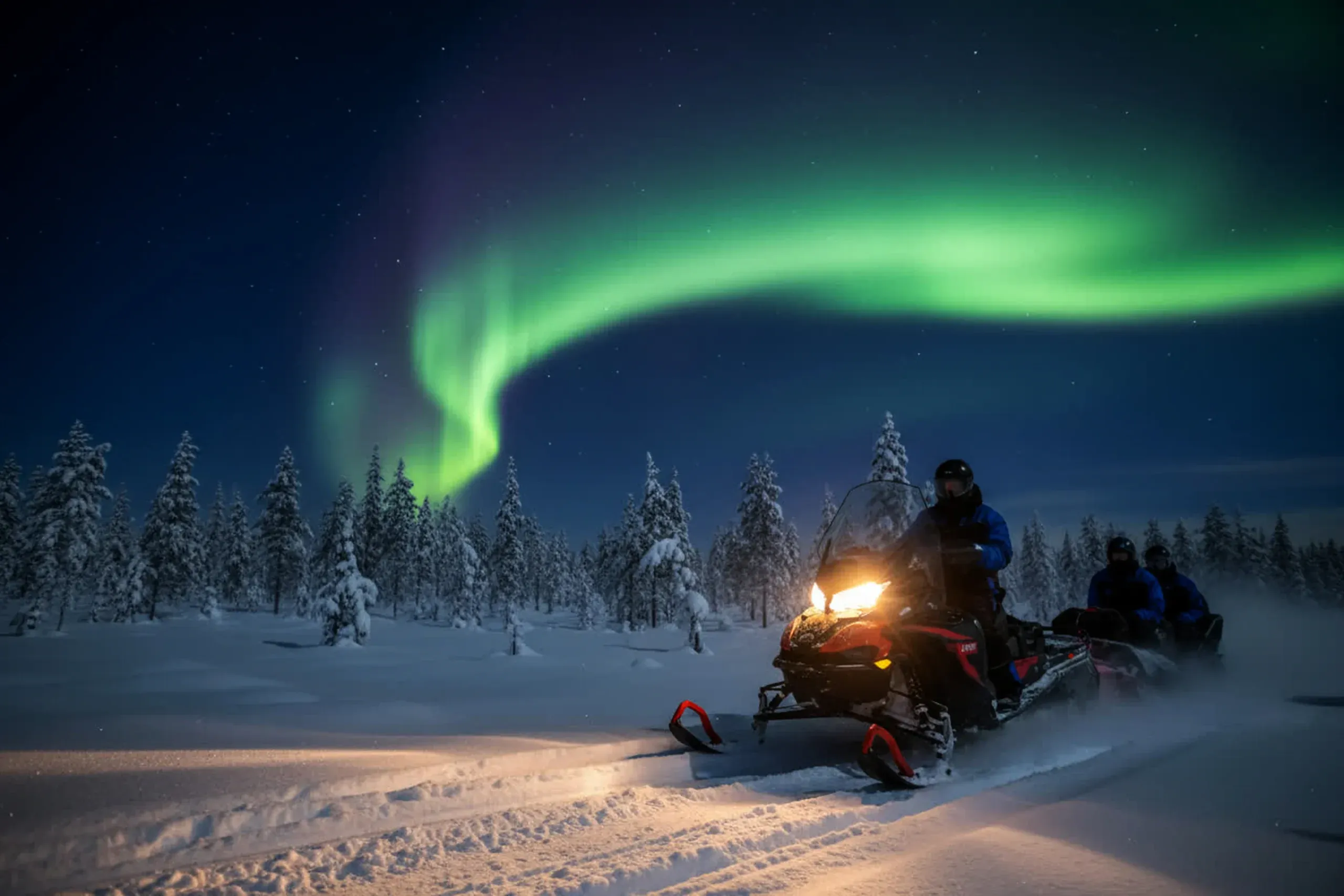 A green aurora arc curving across a dark blue sky above snow-laden spruce forest, with a guide on a snowmobile and two riders on a trail below